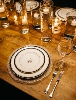 A place setting of gold-rimmed china and cut glass on a bare wooden table, lit by candles.