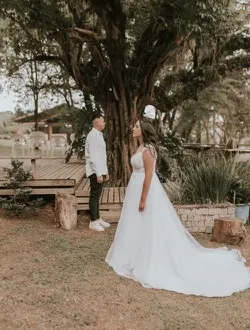 A couple stand together under a large old tree, the bride's dress trailing on the grass.