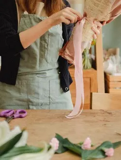 A florist in an apron ties a pale pink ribbon around a wrapped bouquet.