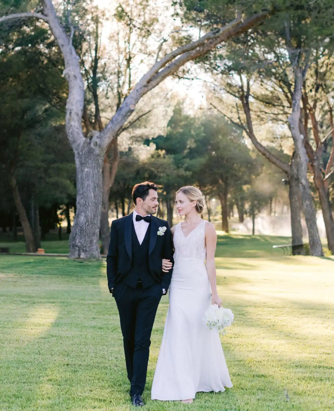 A couple walk arm in arm across parkland in the evening light, the bride carrying white flowers.