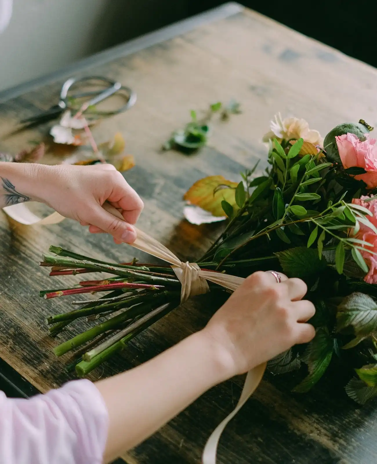 A florist's hands tie a ribbon around the stems of a bridal bouquet on a wooden bench.